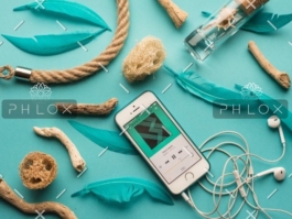 Smartphone playing music surrounded by driftwood pieces, turquoise feathers, and white earphones on a teal background