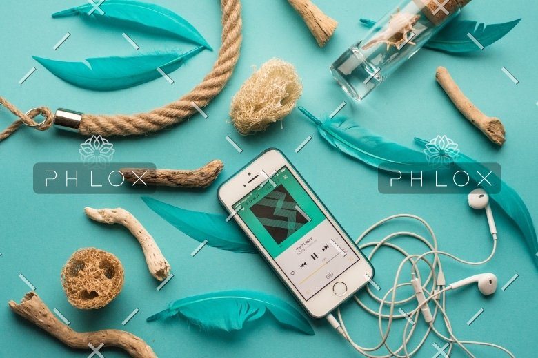 Smartphone playing music surrounded by driftwood pieces, turquoise feathers, and white earphones on a teal background