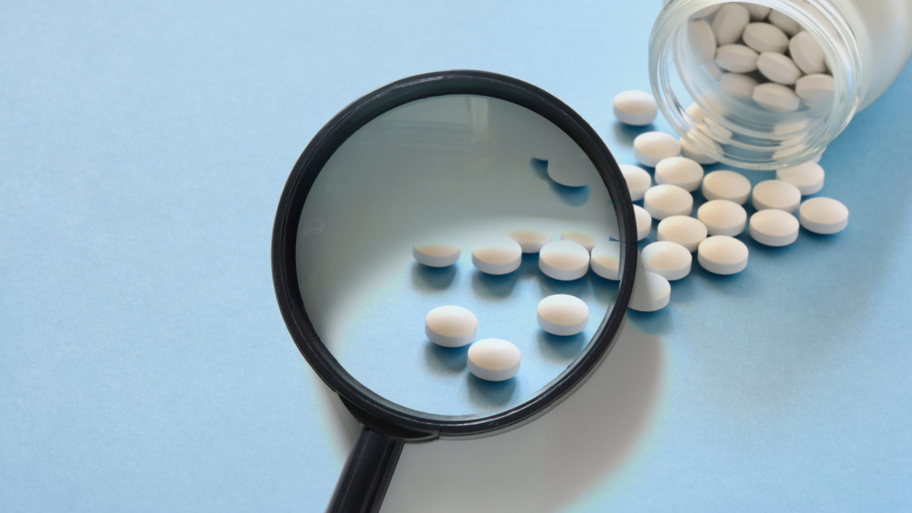 Close-up of white round pills spilled from a bottle with some pills magnified through a black magnifying glass on a light blue background