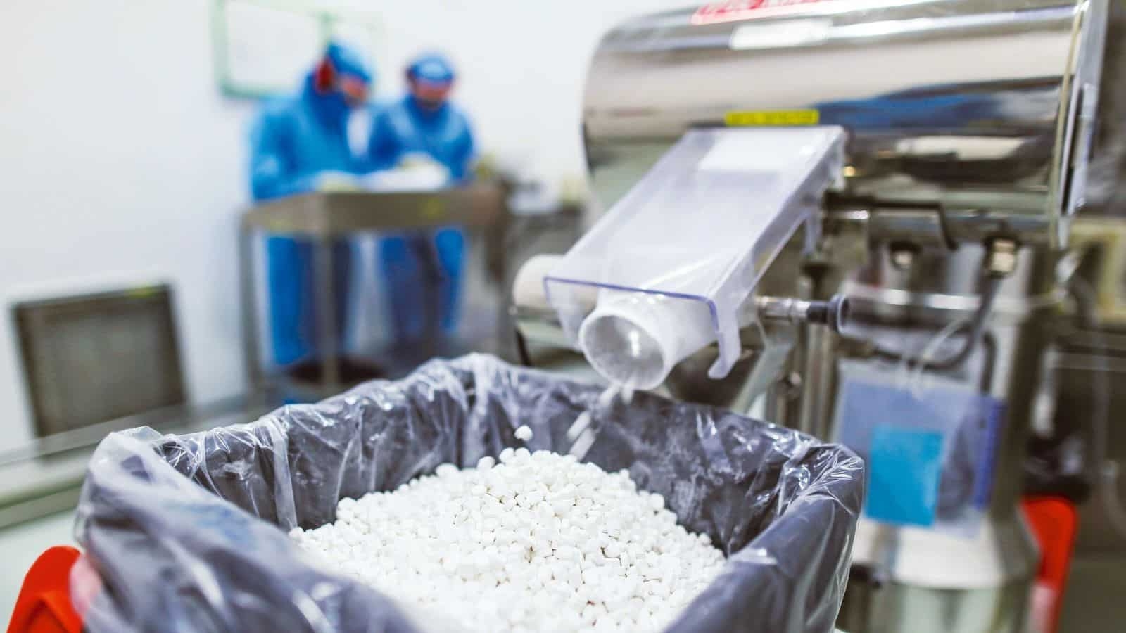 Pharmaceutical manufacturing process with white tablets pouring into a bin and workers in protective gear in the background