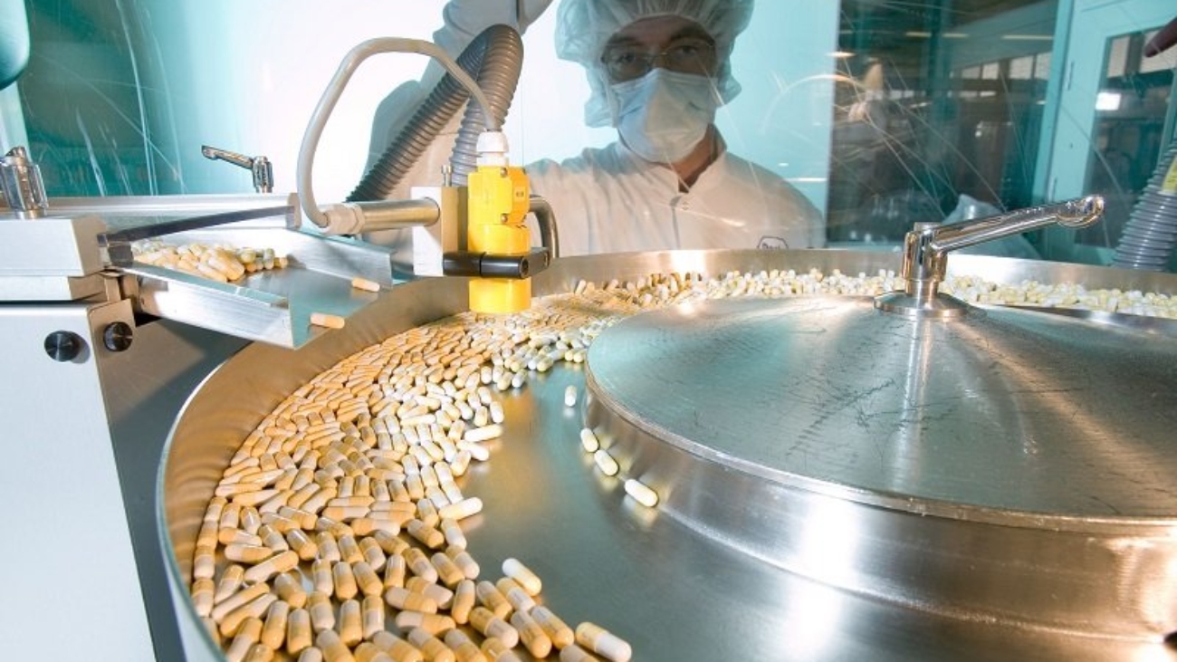Pharmaceutical manufacturing process showing capsules moving on a production line with a worker in protective gear observing