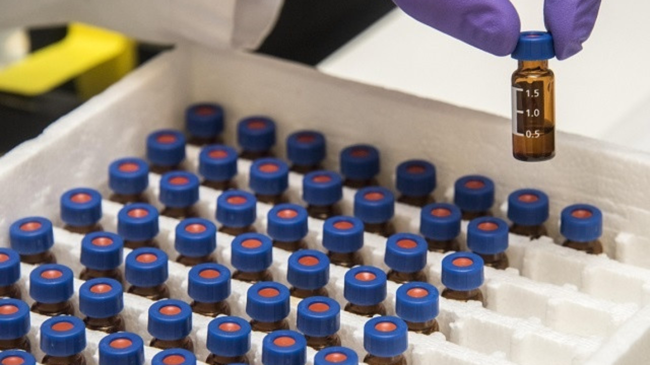 Gloved hand holding a small vial with blue cap above a tray filled with similar vials, representing a peptide or pharmaceutical manufacturer