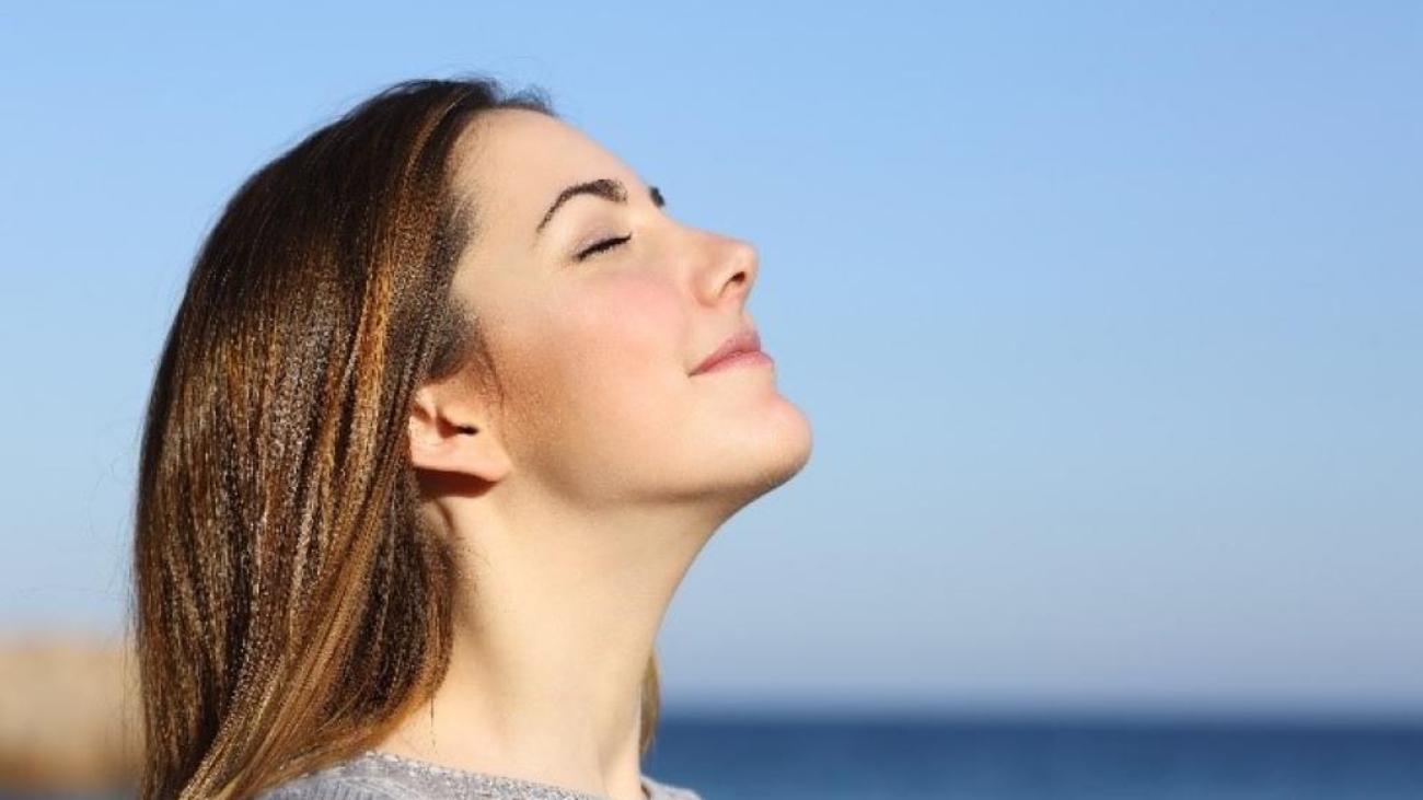 Woman enjoying fresh air with eyes closed by the ocean under clear blue sky