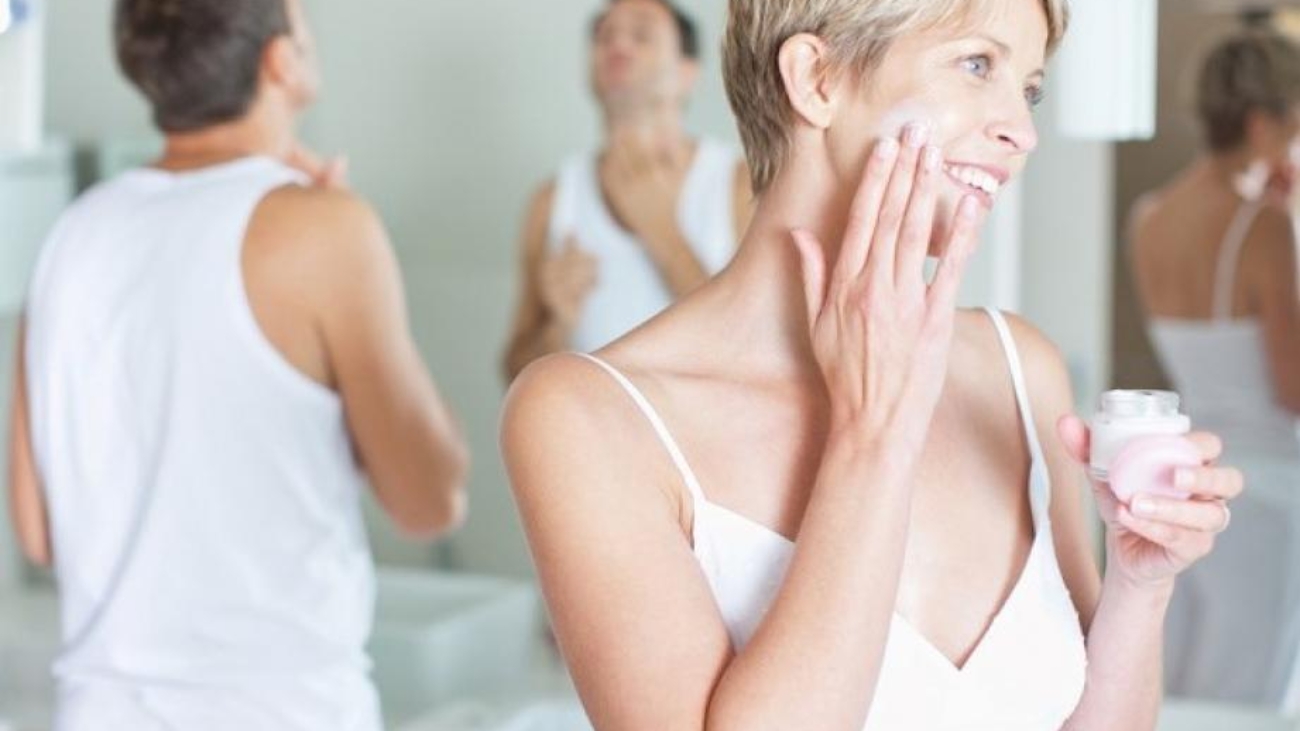 Woman applying face cream in bathroom mirror with man in background