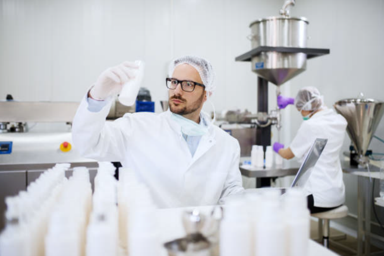 Pharmaceutical manufacturing lab technician inspecting white powder sample in bottle amid automated filling and packaging equipment